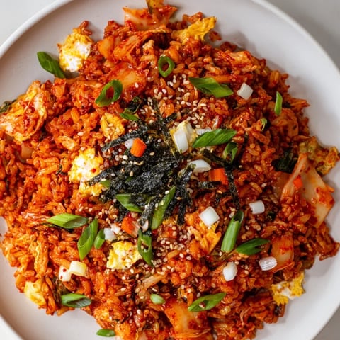 A bowl of steaming Spicy Kimchi Fried Rice, featuring bright red kimchi chunks, fluffy grains, and a fried egg on top with sesame seeds and green onions.  