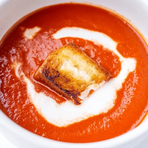 Steaming roasted tomato soup is served in a white bowl, topped with golden grilled cheese croutons and fresh thyme.