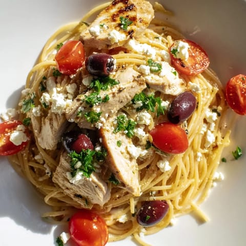 Greek Chicken Spaghetti served hot with al dente pasta, vibrant red cherry tomatoes, and briny olives on a rustic dinner table setting.