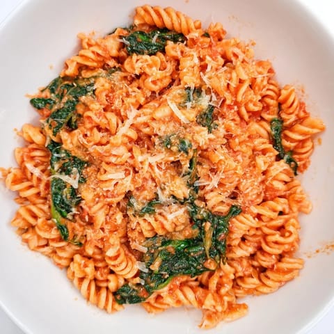 A rustic wooden table holds a pot of freshly cooked Tomato Spinach One-Pot Rotini, garnished with melted Parmesan and fresh basil.