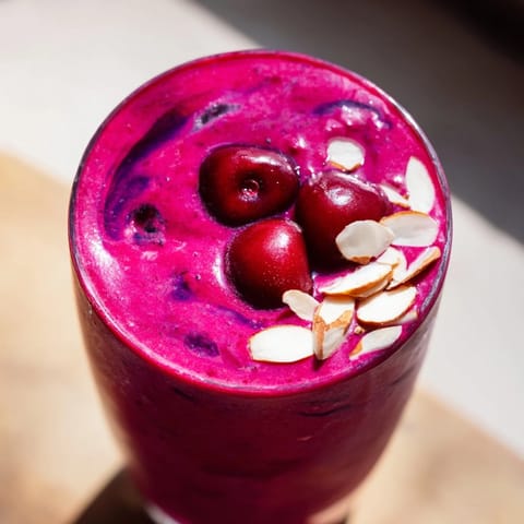 Two frothy glasses of Cherry Almond Smoothie next to fresh cherries and almond butter on a marble counter.  